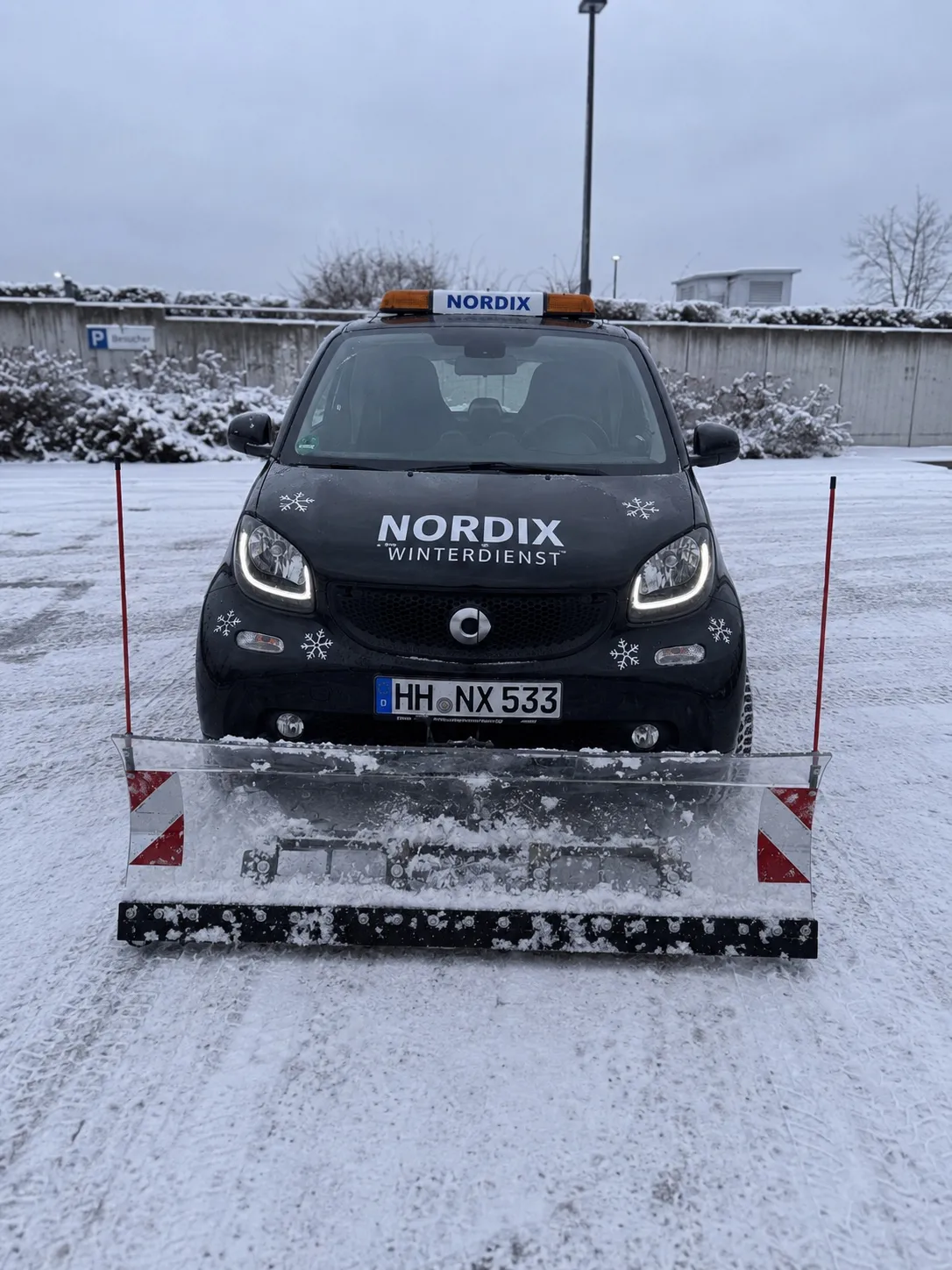 Schwarzer Smart Fortwo von Nordix Winterdienst mit Kennzeichen HH·NX 533, Räumschild und Rundumleuchte auf einem geräumten, verschneiten Parkplatz.