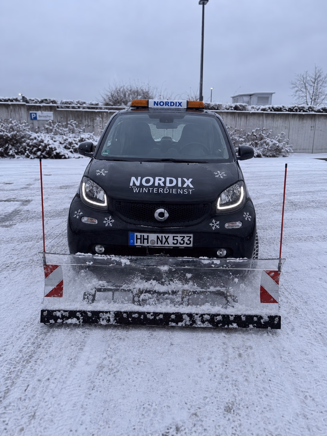 Schwarzer Smart Fortwo von Nordix Winterdienst mit Kennzeichen HH·NX 533 und Räumschild auf einem geräumten, verschneiten Parkplatz.