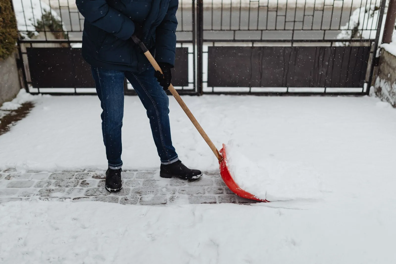 Person in Winterjacke schippt mit einer Schneeschaufel Schnee von einem gepflasterten Gehweg.